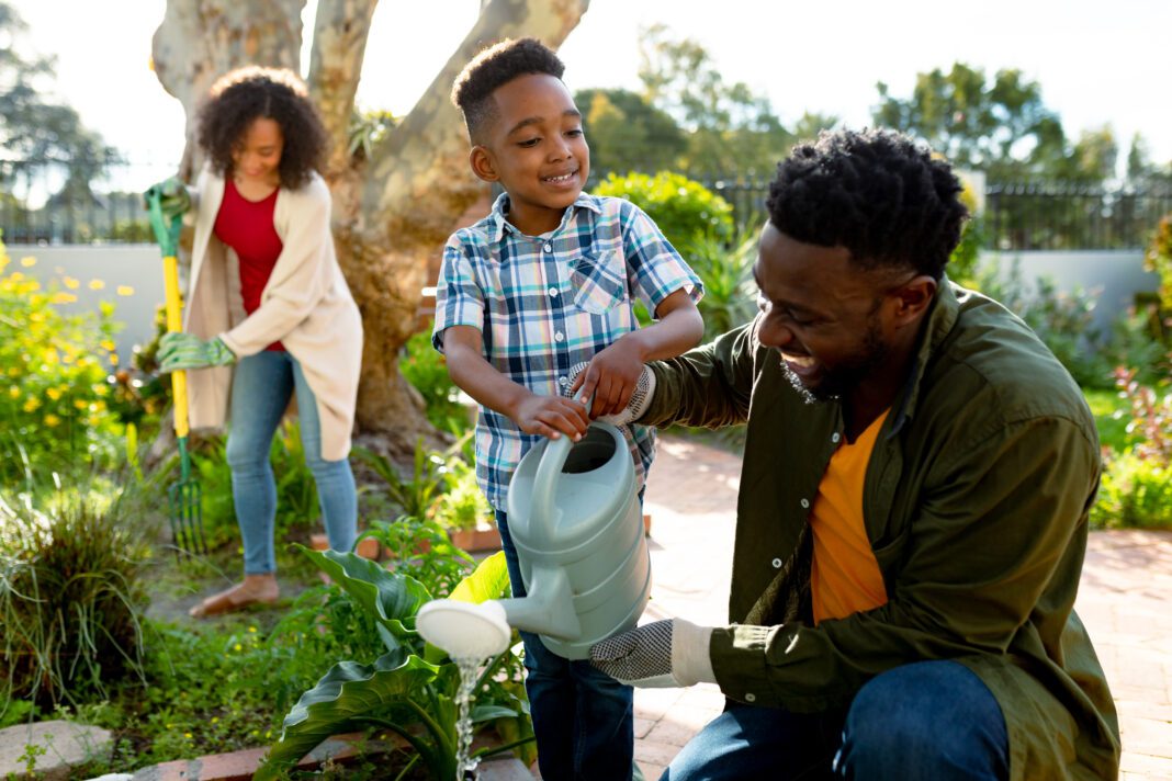 Happy african american father and son watering plants together. family time, having fun together at home and garden.