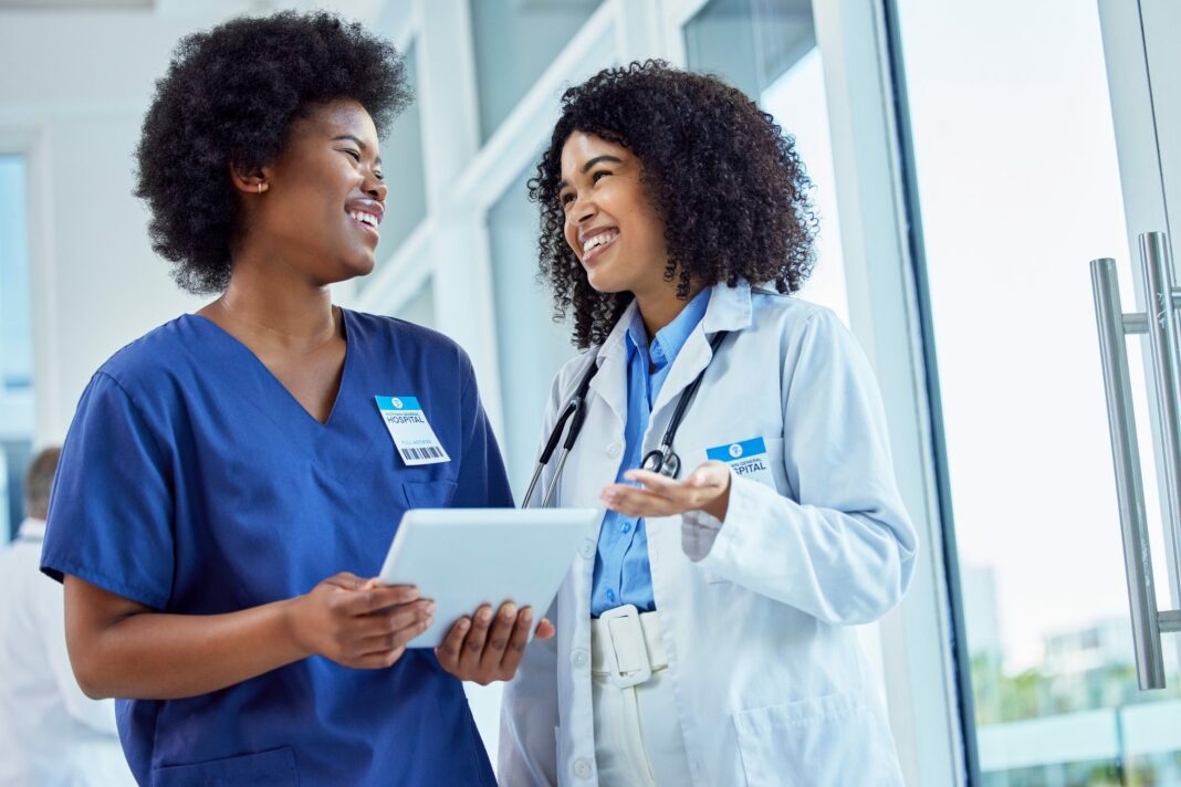 Two black women in nurse coats laugh and look at a tablet