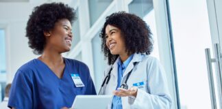 Two black women in nurse coats laugh and look at a tablet