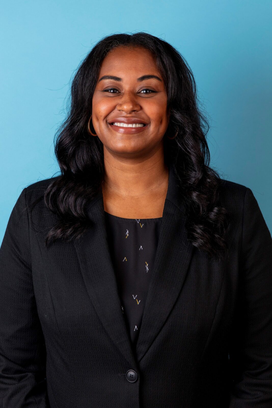 Black woman with dark hair and dark clothing smiles with a blue backround