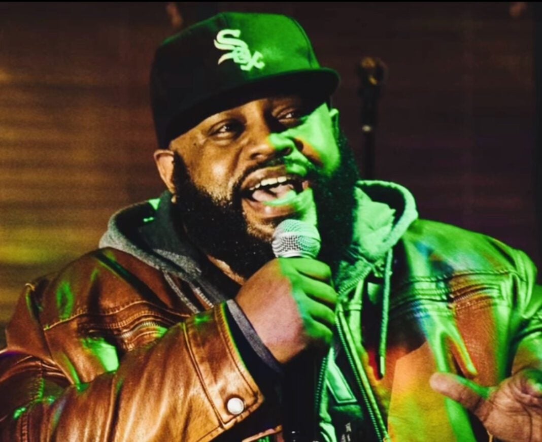 A man in a hoodie, jacket and baseball cap speaks into a microphone