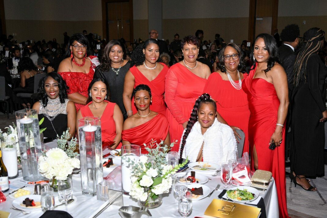 Several women dressed in red pose at a banquet table at a gala