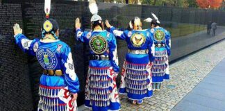 four women in native american jingle dresses stand and touch a vietnam memorial