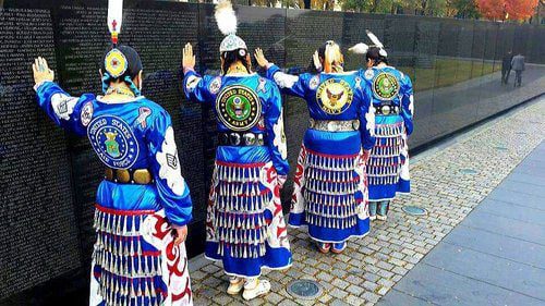 four women in native american jingle dresses stand and touch a vietnam memorial