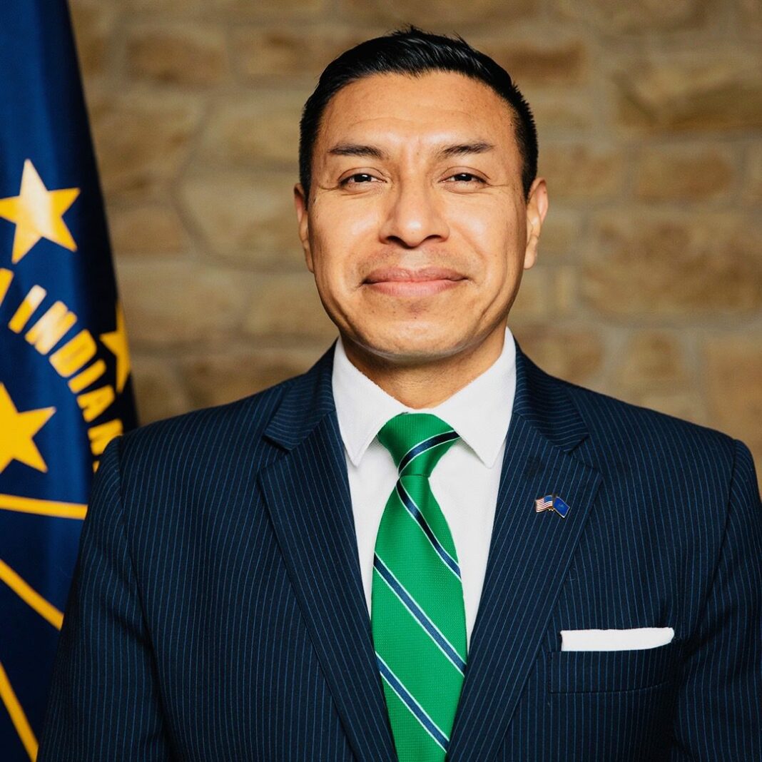 Secretary of State Daniel Morales looking at the camera in a green and blue striped tie and a blue suit with a flag behind him