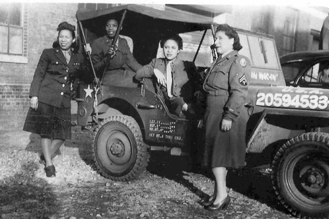 four black women in army uniforms stand in front of a truck