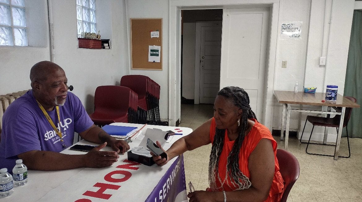 A man in a purple shirt sits at a white table and hands a phone to a lady in an orange dress