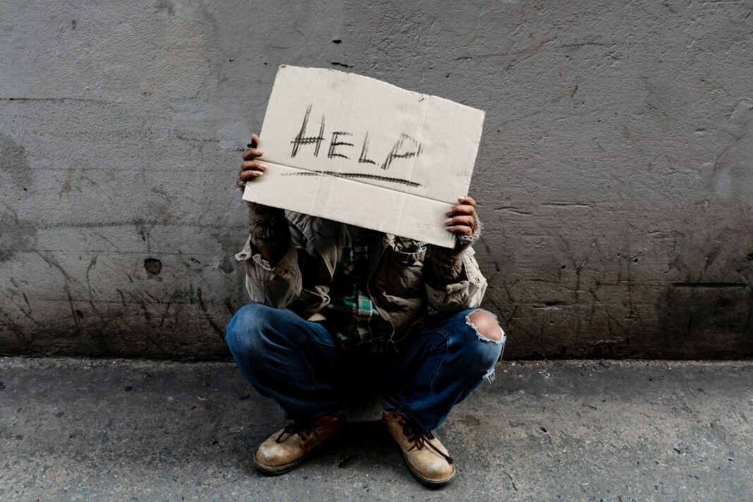 A homeless, long-haired Asian man sits hopelessly leaning against a wall as there is no one to help him with work and food in his hand holding a sign for help. Homeless sleep on streets