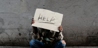 A homeless, long-haired Asian man sits hopelessly leaning against a wall as there is no one to help him with work and food in his hand holding a sign for help. Homeless sleep on streets