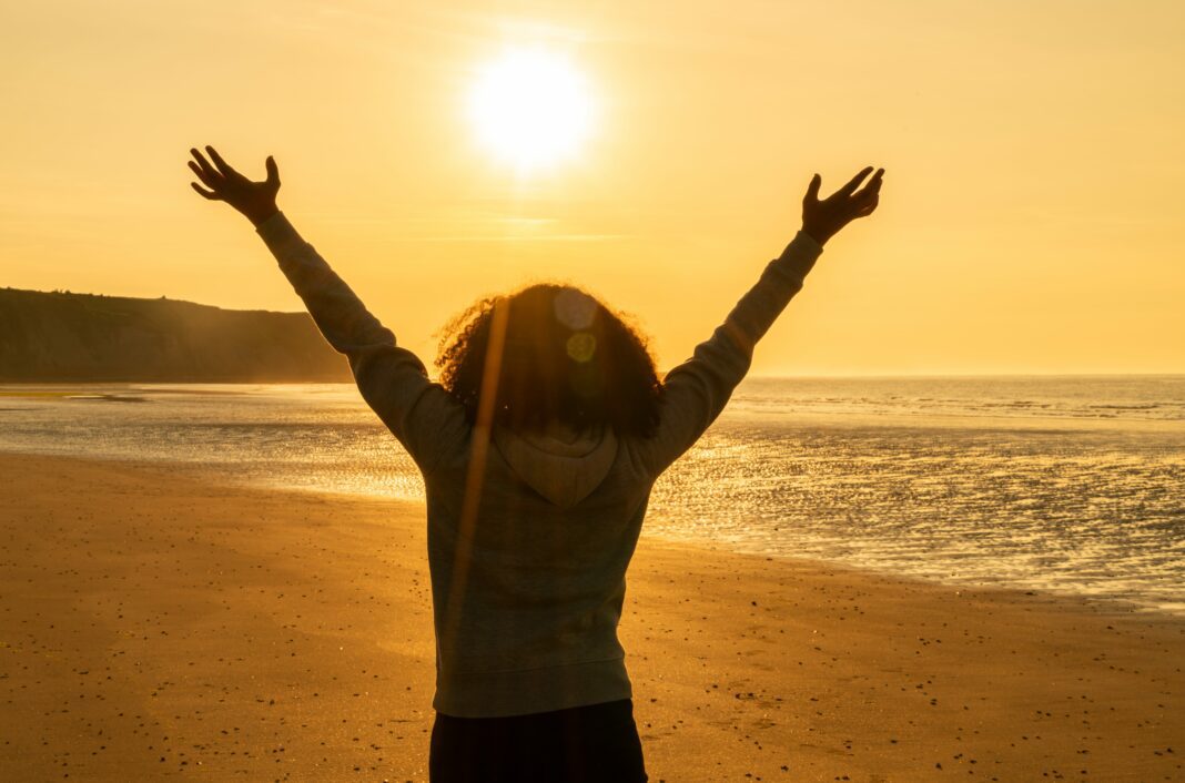 Rear view of young woman girl female standing arms raised in celebration standing on a deserted beach at sunset