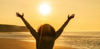 Rear view of young woman girl female standing arms raised in celebration standing on a deserted beach at sunset