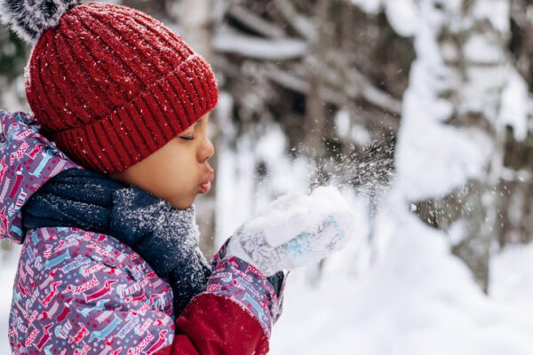 Happy little African-American girl in a red hat and overalls blowing snow off the hand.Winter, Christmas and Happy New Year concept.Copy space.