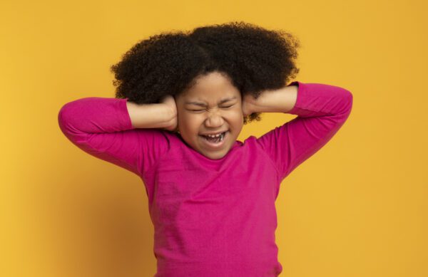 Annoyed little girl covering ears with hands, doesn't want to hear or know something, emotionally reacting to arguing, yellow background