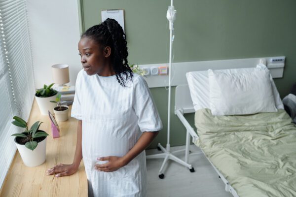 Pregnant woman standing by windowsill while holding her belly in hospital room, gazing outside with peaceful expression. Hospital bed and indoor plants visible in calm environment