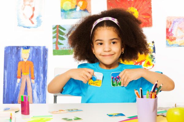 Cute girl playing developmental game holding cards matching relation by table sitting in playroom with wall behind full of children drawings