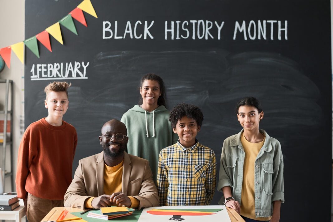 Portrait of African American teacher smiling at camera together with his children while they standing against the blackboard