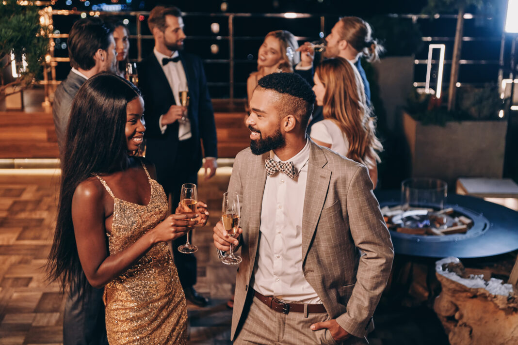 Beautiful young African couple in formalwear holding champagne flutes and smiling while spending time with friends on night party. (Photo/Getty Images)