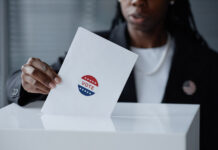 Cropped shot of Black female election participant in business suit casting ballot into voting box, holding envelope with vote sticker at polling station
