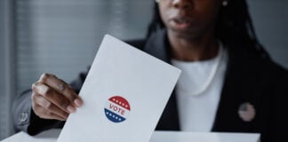 Cropped shot of Black female election participant in business suit casting ballot into voting box, holding envelope with vote sticker at polling station